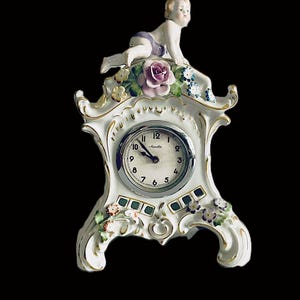 May include: A white porcelain mantel clock featuring a cherub figure on top, a central round clock face with black numerals, and decorative hand-painted floral accents in pink, blue, and green with gold trim along the edges of the clock frame.