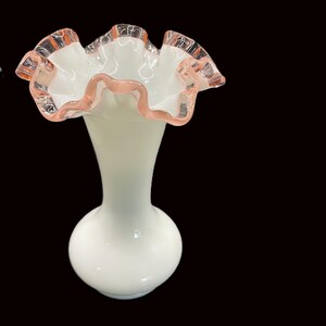May include: A white glass vase with a ruffled rim. The rim has a pink trim. The vase has a bulbous base and a slender neck. The vase is approximately 8 inches tall.