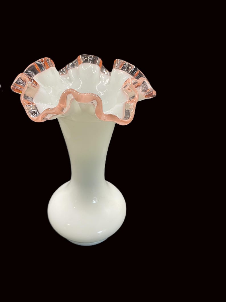 May include: A white glass vase with a ruffled rim. The rim has a pink trim. The vase has a bulbous base and a slender neck. The vase is approximately 8 inches tall.