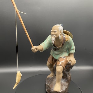 Peut inclure: Figurine en céramique d'un pêcheur âgé assis, tenant une canne à pêche avec un poisson. La figurine porte une chemise verte, un short marron et un chapeau de paille. La figurine est montée sur un socle en forme de rocher brun.
