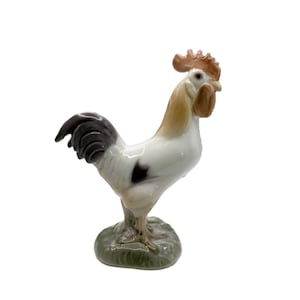 May include: A porcelain rooster figurine standing on a textured green base. The rooster features a white body with brown neck feathers, a dark brown tail, and a prominent red comb and wattle. The ceramic surface has a glossy finish throughout.