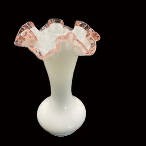 May include: A white glass vase with a ruffled rim. The rim is edged with a delicate pink color. The vase has a bulbous base and a slender neck, creating an elegant silhouette. The background is black.
