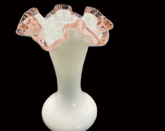 May include: A white glass vase with a ruffled rim. The rim is edged with a delicate pink color. The vase has a bulbous base and a slender neck, creating an elegant silhouette. The background is black.