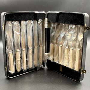 May include: A vintage cutlery set presented in a black case. The set contains knives and forks with cream-coloured handles, each piece individually wrapped in plastic. The case is open, revealing the contents, and has a black velvet lining.