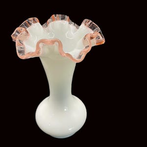 May include: A white glass vase with a ruffled rim, accented with a delicate pink trim. The vase has a bulbous base and a slender neck, creating an elegant silhouette. The ruffled edge adds a touch of vintage charm to this decorative piece.