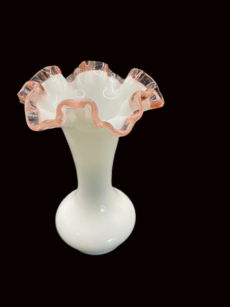 May include: A white glass vase with a ruffled rim, accented with a delicate pink trim. The vase has a bulbous base and a slender neck, creating an elegant silhouette. The ruffled edge adds a touch of vintage charm to this decorative piece.