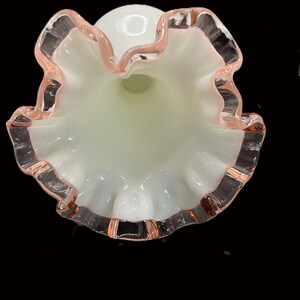 May include: A white glass vase with a ruffled rim, accented with a delicate pink trim. The vase has a flared opening and a smooth, rounded body. The interior has a soft, pale green hue.