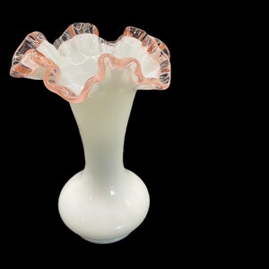 May include: A white glass vase with a ruffled rim. The rim is edged with a delicate pink color. The vase has a bulbous base and a slender neck, creating an elegant silhouette.