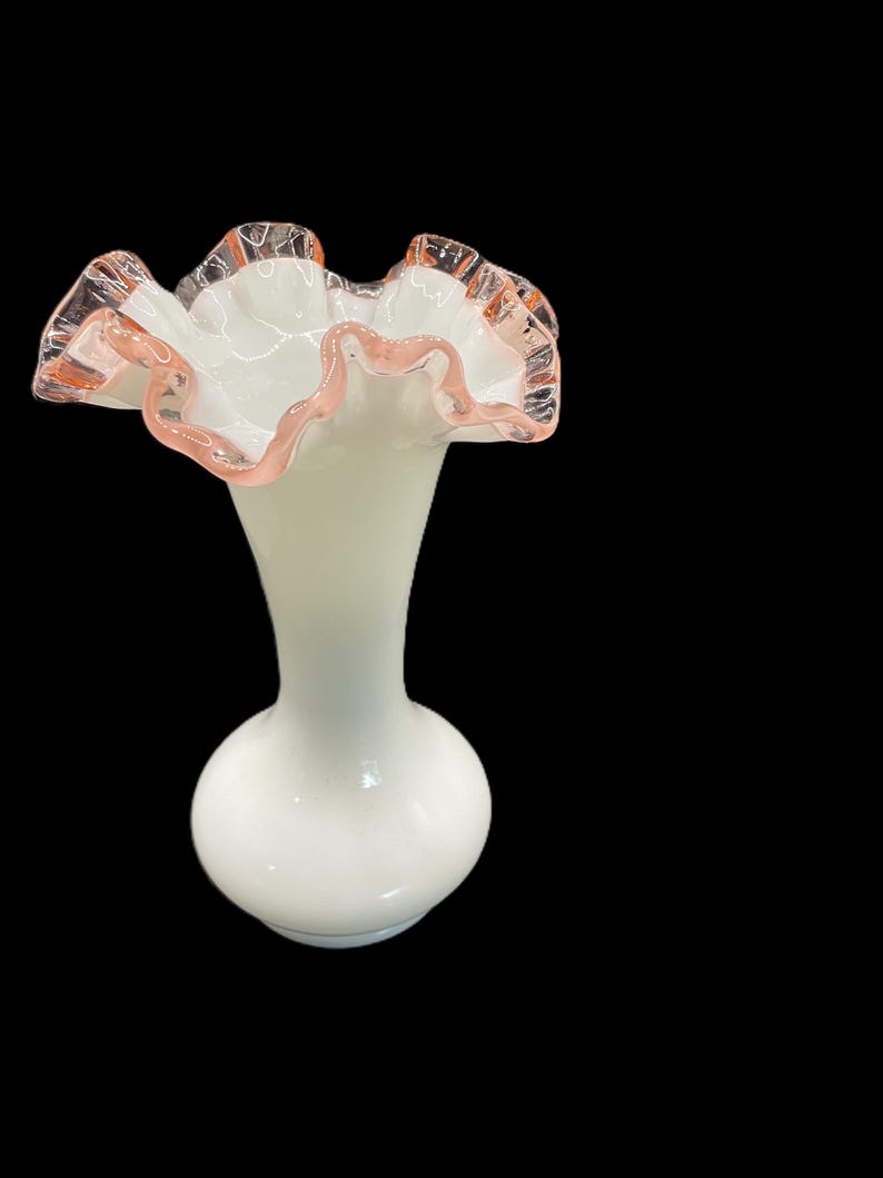 May include: A white glass vase with a ruffled rim. The rim is edged with a delicate pink color. The vase has a bulbous base and a slender neck, creating an elegant silhouette.