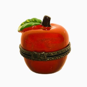 May include: A glossy red apple-shaped trinket box with a dark brown stem and a single green leaf on top. The box features a hinged opening secured by a decorative antique brass-toned metal band with a small bow detail at the front centre.
