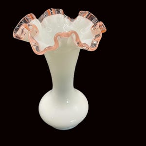 May include: A white glass vase with a ruffled rim. The rim is edged with a delicate pink color. The vase has a bulbous base and a slender neck, creating an elegant silhouette. The vase is likely used for decorative purposes.