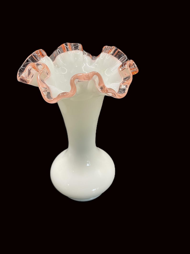 May include: A white glass vase with a ruffled rim. The rim is edged with a delicate pink color. The vase has a bulbous base and a slender neck, creating an elegant silhouette. The vase is likely used for decorative purposes.