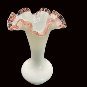 May include: A white glass vase with a ruffled rim and a bulbous base. The rim is trimmed with a delicate pink hue, adding a touch of color to the otherwise monochromatic design. The vase has a slender neck.