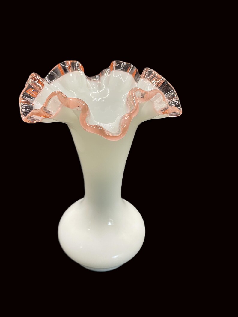 May include: A white glass vase with a ruffled rim and a bulbous base. The rim is trimmed with a delicate pink hue, adding a touch of color to the otherwise monochromatic design. The vase has a slender neck.