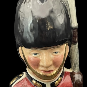 May include: A ceramic figurine depicting a British Royal Guard. It features a black bearskin hat, a red jacket with gold detailing, and a rifle. The face is detailed with blue eyes and a serious expression. The background is black.