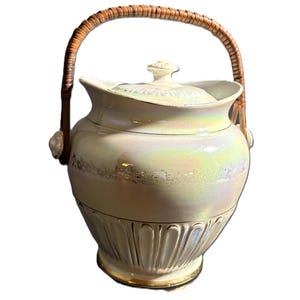 May include: Cream-coloured ceramic serving dish with a lid and a woven handle. The dish has a pearlescent finish and gold trim. The handle is made of woven brown material. The dish has a decorative, fluted base.