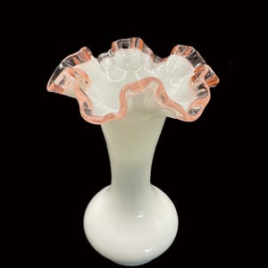 May include: A white glass vase with a ruffled rim. The rim is edged with a delicate pink color. The vase has a bulbous base and a slender neck, creating an elegant silhouette. The vase is likely for decorative purposes.
