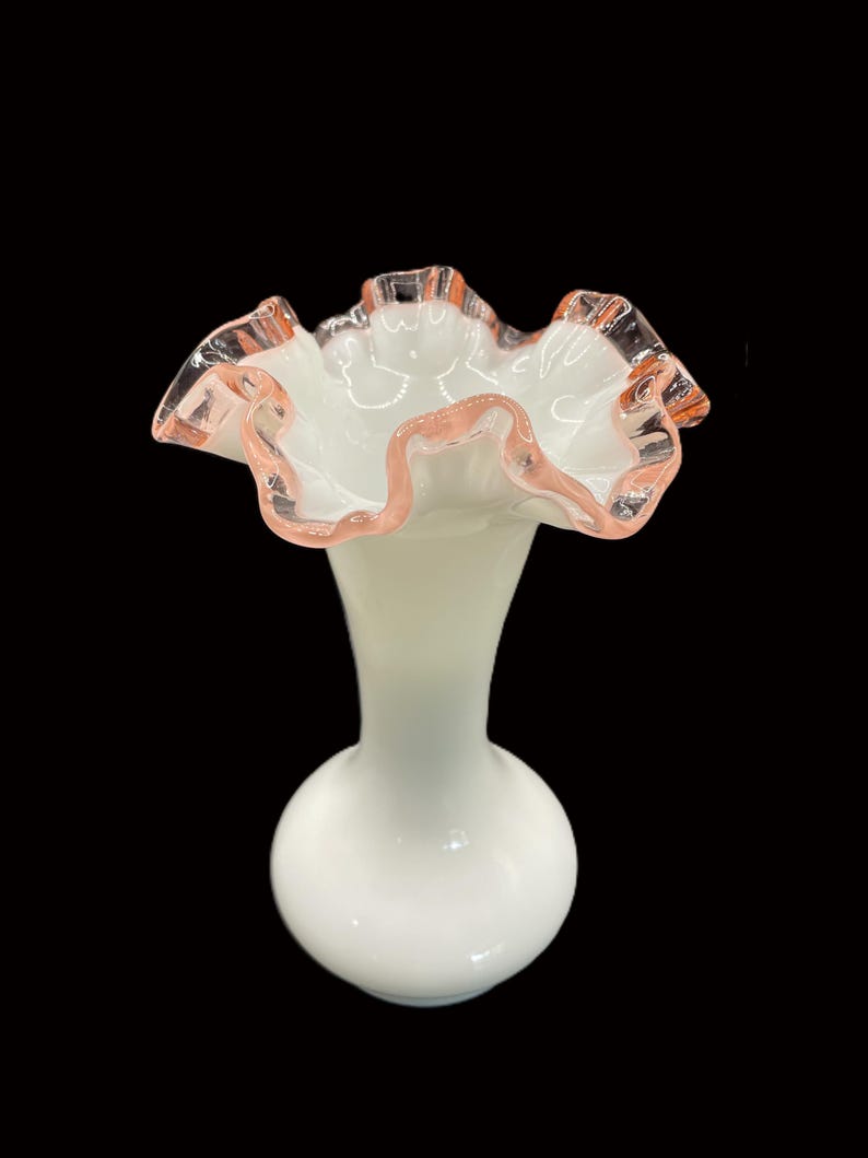 May include: A white glass vase with a ruffled rim. The rim is edged with a delicate pink color. The vase has a bulbous base and a slender neck, creating an elegant silhouette. The vase is likely for decorative purposes.
