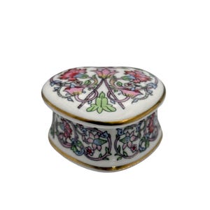 May include: A heart-shaped porcelain trinket box with a white base and gold-trimmed edges. The lid and sides feature a colorful floral pattern with pink, red, green, and blue botanical motifs, including stylized flowers and leaves on a white surface.
