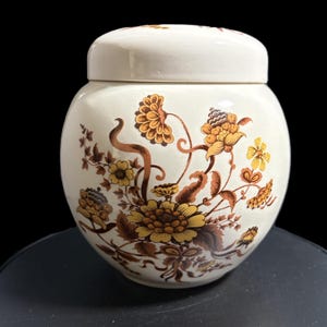 May include: Cream-colored ceramic jar with a matching lid. The jar is adorned with a floral pattern in shades of yellow, brown, and gold. The jar has a round shape and is likely used for storage.