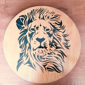 May include: A round wooden wall art piece featuring a lion's head design. The lion's face is cut out of the light-colored wood, revealing a dark teal background. The lion's mane and facial features are intricately detailed.