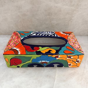 May include: A rectangular ceramic tissue box cover featuring a vibrant, hand-painted floral and geometric pattern in shades of orange, blue, green, red, and yellow. The top surface has a central oval opening with a dark blue rim.