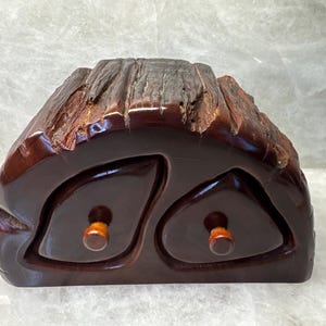 May include: A handcrafted, dark brown wooden box with two drawers, each featuring a small, round, wooden knob. The top of the box has a textured, bark-like appearance, adding a rustic aesthetic. The box is likely used for storing small items.