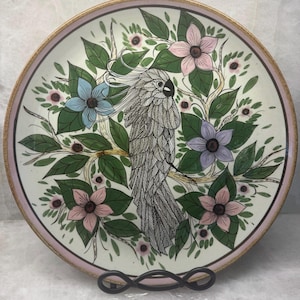 May include: A decorative ceramic plate featuring a detailed illustration of a cockatoo bird surrounded by flowers and foliage. The plate has a cream-colored background with a pink and gold rim. The bird is primarily gray and white, with accents of blue, pink, and purple.
