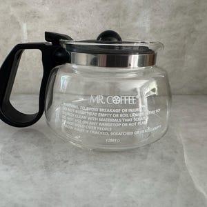 May include: Clear glass Mr. Coffee coffee pot with a black handle and lid. The pot has a silver rim and the Mr. Coffee logo is visible. Text on the pot warns against misuse. A kitchen appliance.