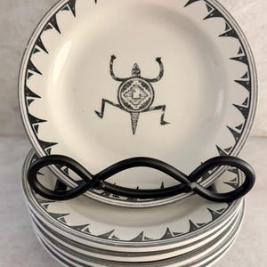 May include: A stack of round ceramic plates in off-white with a black graphic of a lizard-like creature centered on the top plate. The rim of each plate features a repeating black triangular pattern and thin circular bands around the edge.