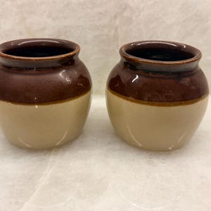 May include: Two small, round ceramic pots featuring a two-tone design. The upper portion of each pot is a glossy dark brown, while the lower portion is a smooth, matte beige. Both containers have a wide, open mouth and a curved, bulbous body shape.