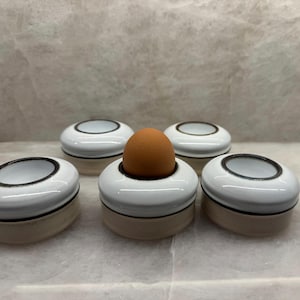 May include: Five white and beige egg cups with a black rim. One egg cup holds a brown egg. The egg cups have a minimalist design and are arranged on a light-colored surface.