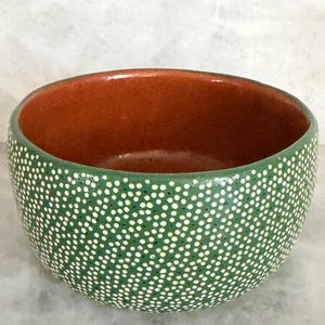 May include: A round ceramic bowl featuring a deep terracotta interior and a vibrant green exterior surface. The outer wall is decorated with a dense, uniform pattern of small, hand-painted white dots covering the entire green surface area.