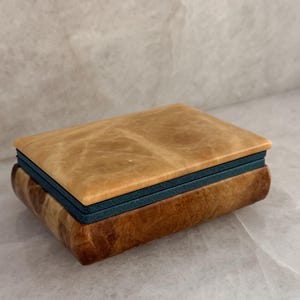 May include: A rectangular box with a smooth, light tan, marbled lid and a dark, textured, brown wooden base. The lid and base are separated by a thin, dark teal-colored layer that runs along the perimeter of the box, creating a distinct contrast.