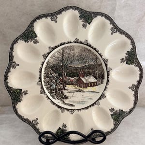 May include: A cream-colored ceramic egg plate featuring twelve oval indentations and a scalloped edge. The border is decorated with a black and green leaf pattern. The center depicts a winter landscape with a church and bare trees.