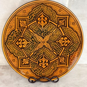 May include: A round ceramic plate featuring a vibrant orange and dark brown geometric pattern. The design includes four symmetrical floral-inspired motifs separated by intricate lines, with stippled green and brown textures filling the gaps.