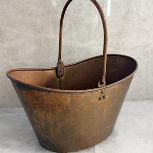 May include: A weathered copper bucket with a wide, oval-shaped opening and a tall, arched metal handle. The exterior surface displays a natural patina with dark, mottled oxidation marks and visible rivets securing the handle to the body.