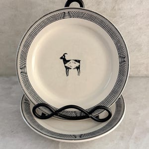 May include: Two round ceramic plates featuring a cream-colored base and a black goat illustration in the center. The rim of each plate is decorated with a black and white grid pattern. The plates are displayed on a black metal wire stand.