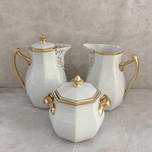 May include: A three-piece white porcelain tea set featuring a teapot, a creamer, and a sugar bowl with a lid. Each piece has a hexagonal body, gold-trimmed edges, and gold-painted handles. The teapot and sugar bowl include matching gold finials.