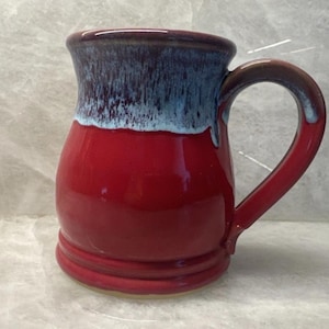 May include: A handmade ceramic mug with a vibrant red base, transitioning to a blue and brown rim. The mug features a curved handle and a glossy finish. The mug is a unique piece of drinkware.