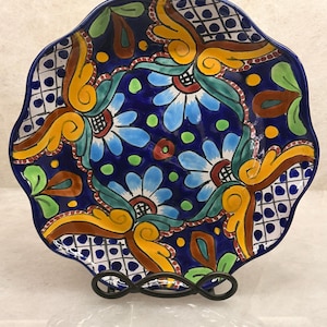 May include: A round ceramic plate with a scalloped edge featuring a vibrant hand-painted pattern. The design includes light blue flowers, yellow scrollwork, green leaves, and blue and white grid sections against a deep blue background.