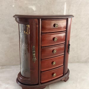 May include: A dark wood jewelry armoire featuring five horizontal drawers with small round brass knobs. The unit includes a side cabinet with a curved glass door etched with floral patterns and a vertical brass handle on the side frame.