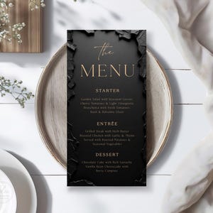 May include: A black menu card with gold lettering, featuring the words "The MENU". The menu lists starter, entree, and dessert options. The card is set on a large, neutral-toned plate, with white flowers and a white cloth in the background.
