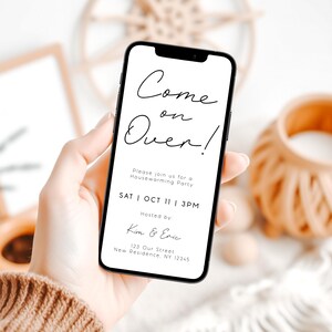 May include: A smartphone displays a housewarming party invitation with the text "Come on Over!" in elegant script. The invitation includes the date, time, and hosts' names. The phone is held in a hand, with a neutral background.