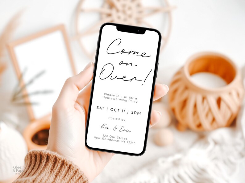 May include: A smartphone displays a housewarming party invitation with the text "Come on Over!" in elegant script. The invitation includes the date, time, and hosts' names. The phone is held in a hand, with a neutral background.