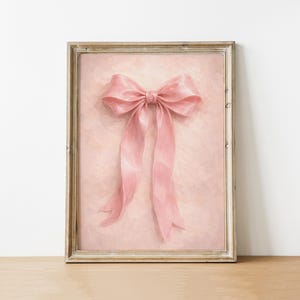 May include: A framed artwork featuring a large, pink bow with long, flowing ribbons. The bow is centered on a textured, light pink background. The frame is a light, neutral color, and the artwork is displayed on a wooden surface.