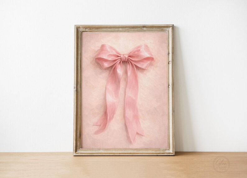 May include: A framed artwork featuring a large, pink bow with long, flowing ribbons. The bow is centered on a textured, light pink background. The frame is a light, neutral color, and the artwork is displayed on a wooden surface.