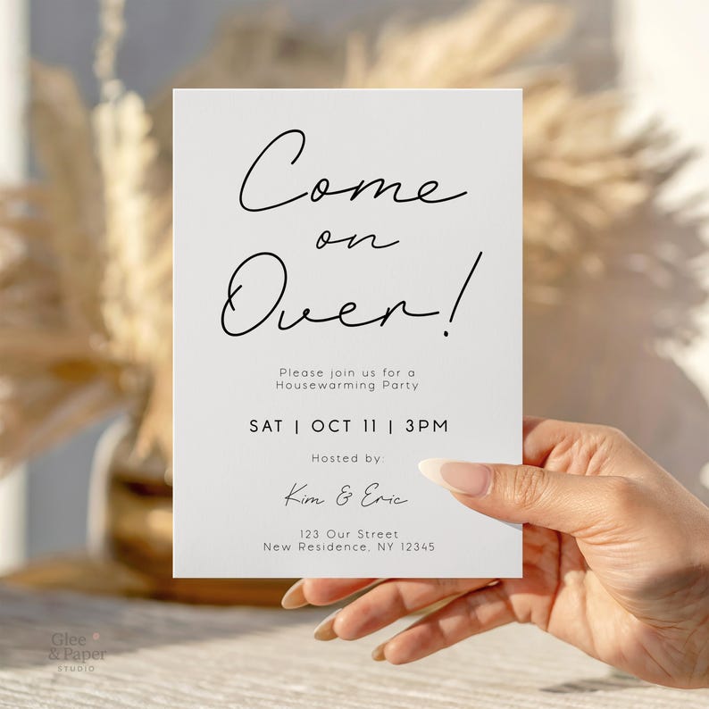 May include: A white invitation card with the words "Come on Over!" in elegant black script. The card also includes details for a housewarming party: date, time, and hosts' names. The card is held by a person.