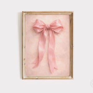 May include: A framed print featuring a large, pink bow with long, flowing ribbons. The bow is centered against a textured, light pink background. The frame is a light, distressed gold color, adding a vintage aesthetic.