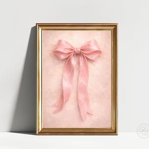 May include: A framed artwork featuring a large, pink bow. The bow is the focal point, set against a soft, textured pink background. The frame is gold-colored, adding a touch of elegance to the piece. The artwork is a decorative item.